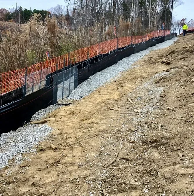 Silt fence installed along the downslope edge of a graded residential construction lot in Charlotte, NC. Orange-staked permeable fabric barrier holds disturbed soil on-site with seed straw applied to the exposed ground surface behind it.
